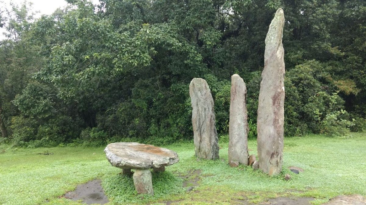 Three standing stones and a flat table stone on a lawn at the edge of a dense forest in Meghalaya.