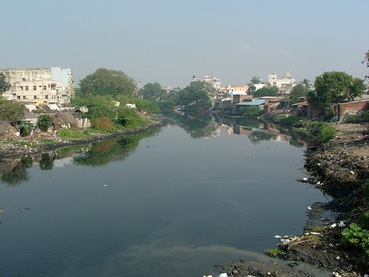 A dark, slow river between banks of rubbish and scrub, with houses on both sides.