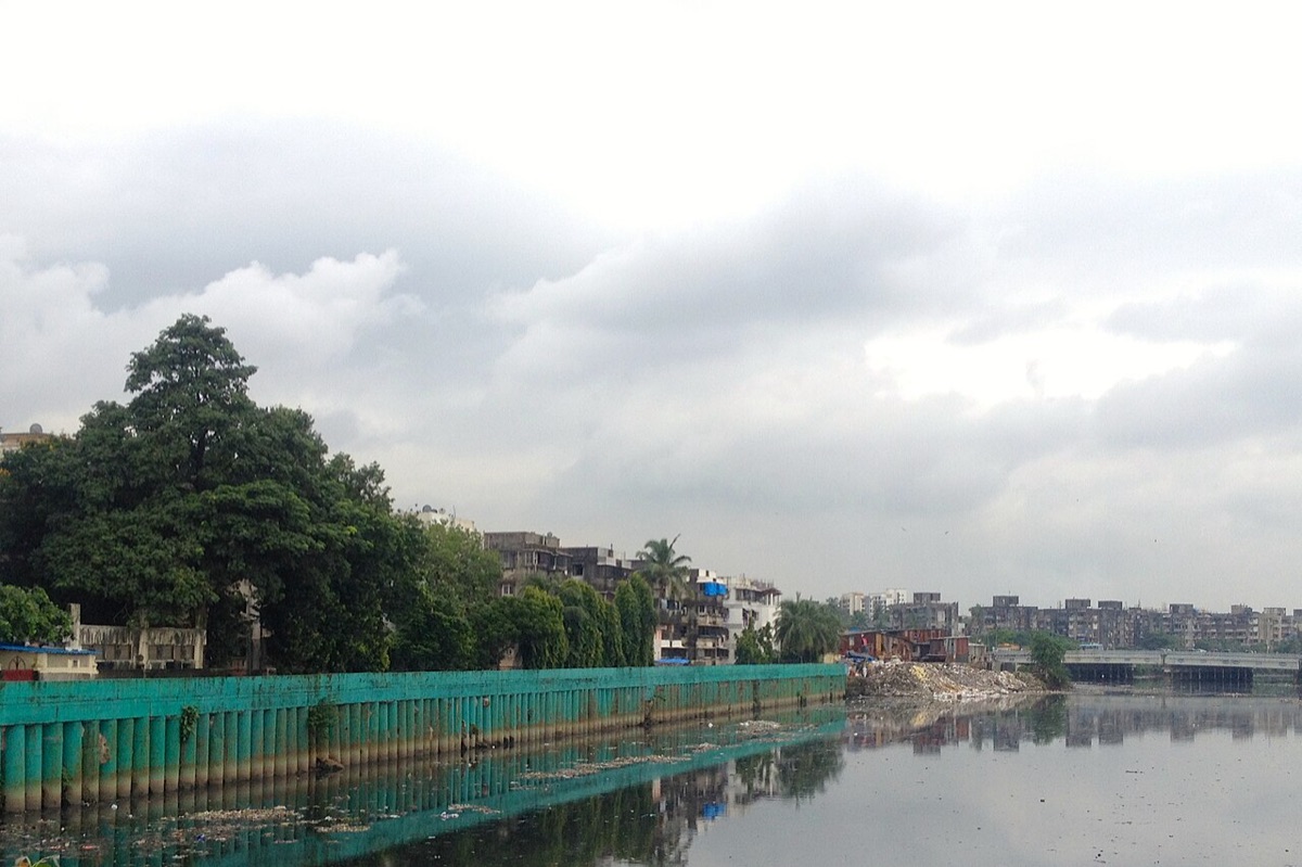 A wide, still, grey-green channel lined with a green wall, trees and low buildings under a cloudy sky.